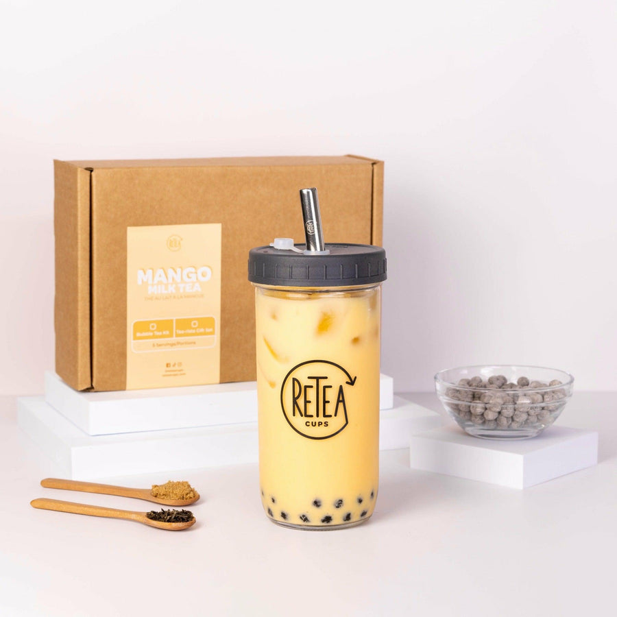 Mango milk tea package box, black Retea reusable bubble tea cup and chrome stainless steel straw, clear glass boba cup containing yellow mango milk tea with brown sugar tapioca, uncooked tapioca boba pearls in a clear bowl, raw dark brown sugar in wooden spoon, jasmine green tea leaves in wooden spoon
