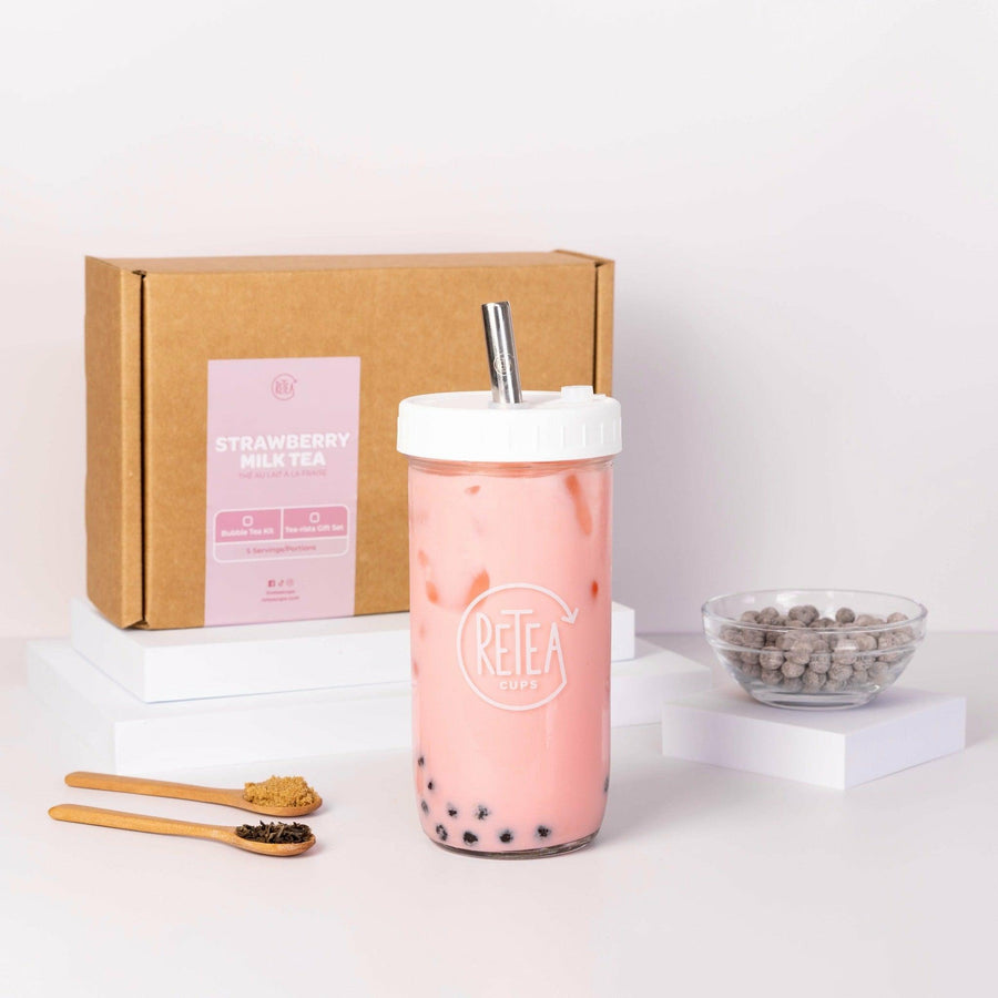 Strawberry milk tea package box, white Retea reusable bubble tea cup and silver reusable boba straw, clear glass bubble tea drink cup containing pink strawberry milk bubble tea with brown sugar tapioca, uncooked tapioca pearls in a clear bowl, raw dark brown sugar in wooden spoon, jasmine green tea leaves in wooden spoon