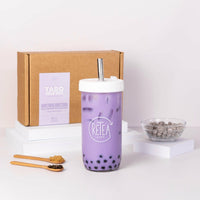 Taro milk tea package box, white Retea reusable bubble tea cup and silver stainless steel straw, the clear glass boba cup contains purple ube milk tea with brown sugar tapioca, uncooked tapioca pearls in a clear bowl, raw dark brown sugar in wooden spoon, jasmine green tea leaves in wooden spoon